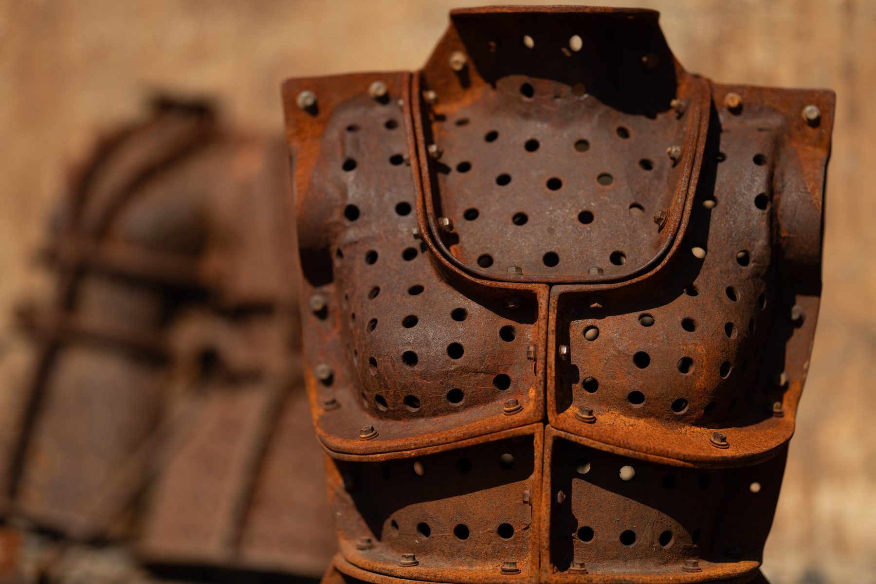A rusted perforated steel torso sculpture with bolted plates, lit by low sun against a soft brown background