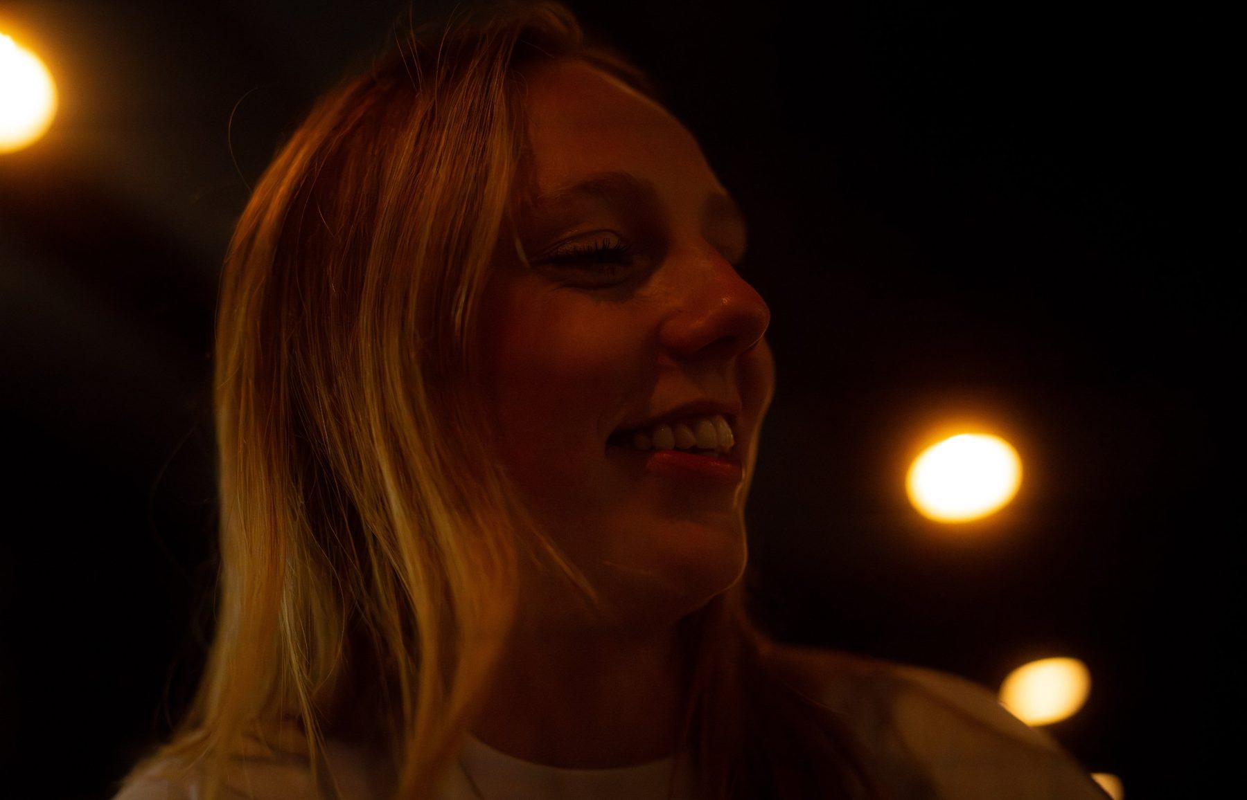 A woman smiling with her eyes closed, her face lit warm amber against a dark room with glowing round lights