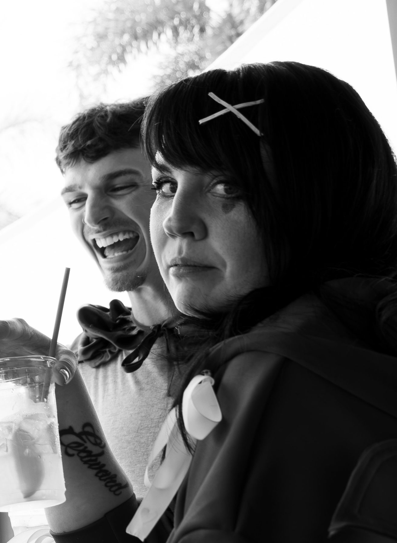 A man laughing with his eyes shut behind a woman who glances at the camera with a dry, knowing look