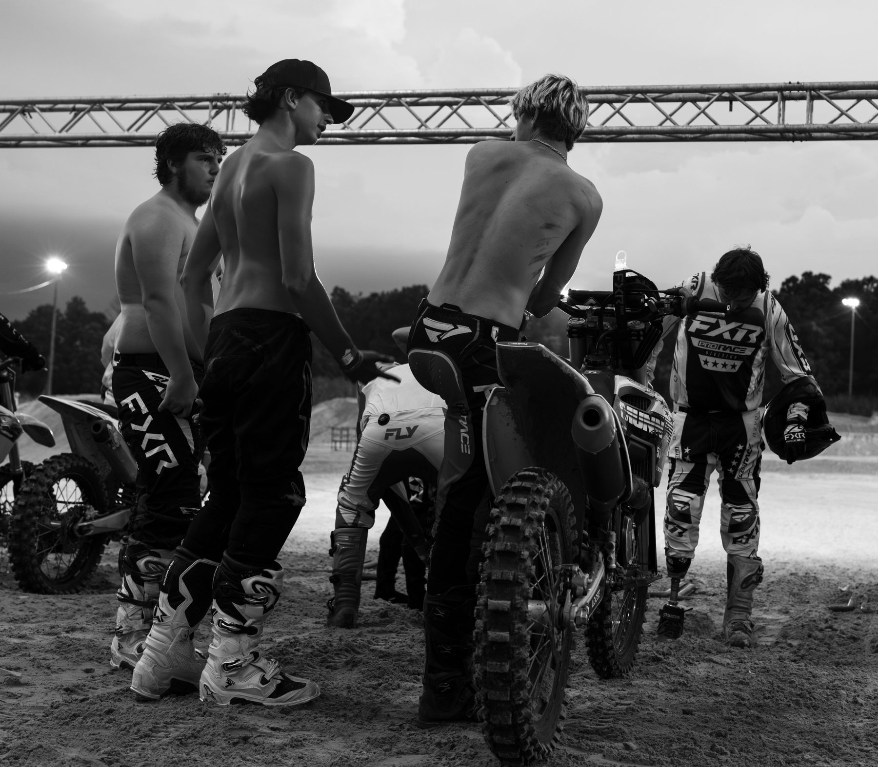 Motocross riders in black and white, shirtless and half dressed in race gear, talking beside their bikes under a steel truss at dusk