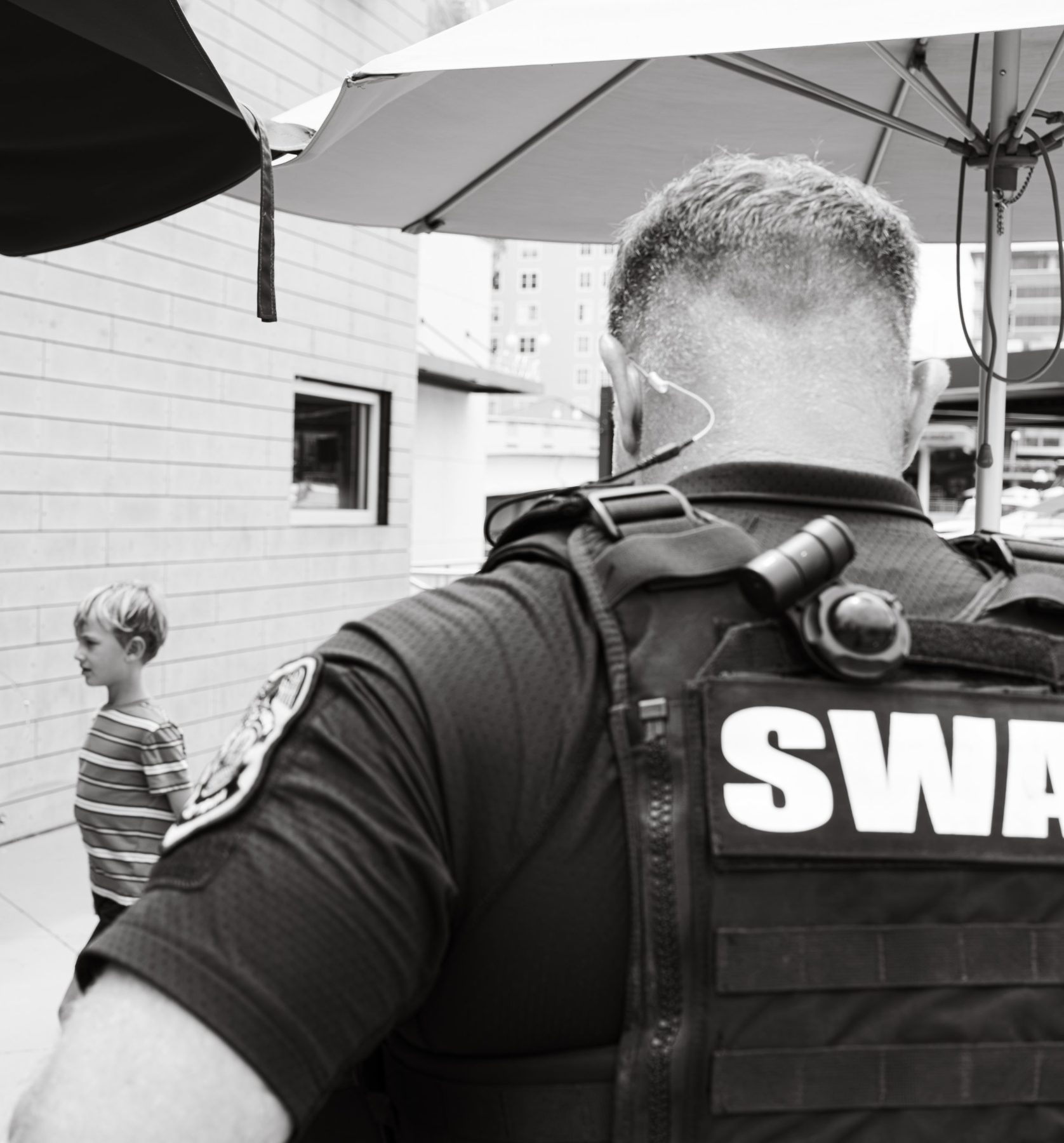 A SWAT officer seen from behind in the foreground while a small boy in a striped shirt looks away in the distance