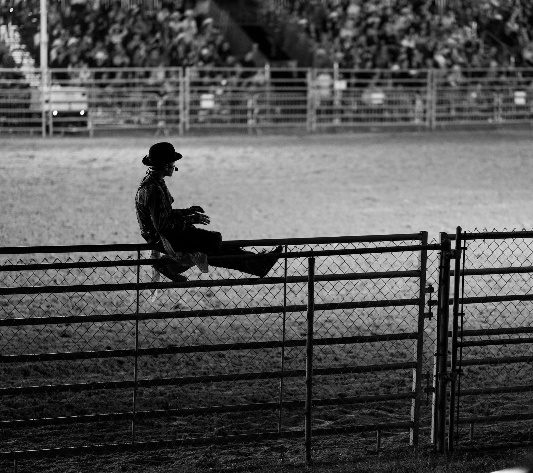 A rodeo clown in a bowler hat sits alone on a pipe fence, watching the empty arena while the crowd blurs behind him